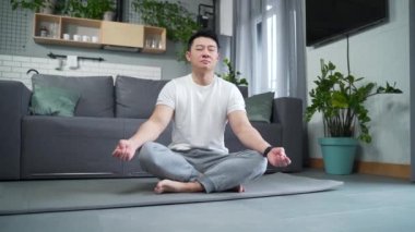 Asyalı sporcu yoga yapıyor. Oturma odasında nilüfer pozisyonunda oturuyor. Sakin erkek meditasyon yapar ve evde dinlenir. Fitness sporu ve sağlıklı aktif yaşam tarzı. Gözleri kapalı nefes alıyor.