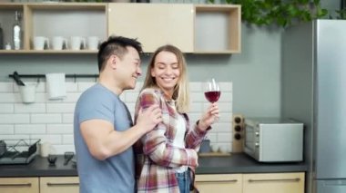 Erkek ve kadın birlikte dans ediyor, evde, mutfakta, mutlu bir ailede eğleniyorlar. Mutlu, çok kültürlü Asyalı çift birlikte vakit geçirip bir kadeh kırmızı şarapla kadeh kaldırıyor.
