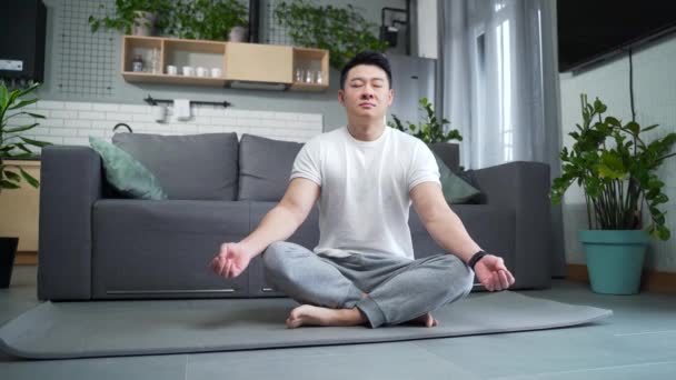 リビングルームの自宅で蓮の位置にマットの上に座ってヨガを練習するアジアのスポーティな男 落ち着いた男性は屋内で瞑想しリラックスします フィットネススポーツと健康的なアクティブライフスタイル 目を閉じて呼吸する — ストック動画