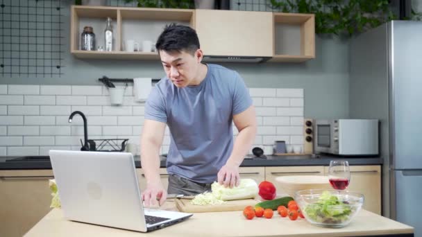 キッチンでビデオチュートリアルの準備をしているアジア人男性 ある男がインターネットで料理のチュートリアルを見ている 家庭のキッチンでノートパソコンを使ってオンラインで料理を作る男性は — ストック動画
