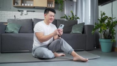 Atletik Asyalı adam evde yoga minderinde oturuyor ve akıllı telefon kullanıyor. Video dersleri izleyin. Yoga, antrenman, egzersiz. Fitness erkeği cep telefonuna bakar. İç mekan Mutlu atlet okur