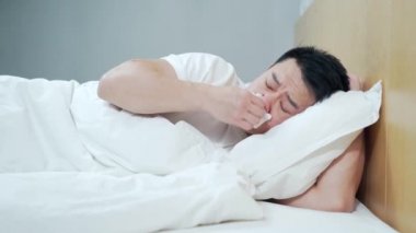 Asyalı bir adam yatakta hasta yatıyor, öksürüğü akıyor ve evde battaniyeyle kaplanmış bir grip. Burnunu peçeteye sümkürdü. Ateşi ve baş ağrısı olan bir erkek. Hastalık. Enfeksiyon, salgın, basil taşıyıcı