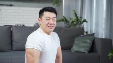 Antrenmandan ya da evde yoga yaptıktan sonra yorgun düşmüş Asyalı bir adamın portresi. İç mekan. Atletik erkek kameraya bakıyor. Terli, kaslı, formalı adam. Neşeli ve mutlu bir şekilde bir paspasın üzerinde oturuyor. 