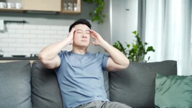 Evde yalnız başına yorgun ve hasta bir adam, Asyalı 'nın başı ağrıyor, kanepede oturuyor ve ellerini başının arkasında tutuyor. Şakaklara masaj yapan hasta bir erkeğin yakın çekimi. Ağrı, acı, kan basıncı