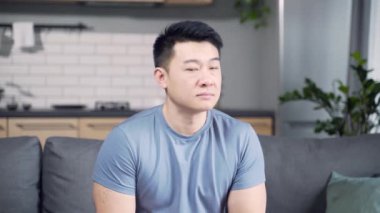 Ev kıyafetleri içinde, bir apartman dairesinde ya da evde kameraya güvenle bakan Asyalı genç bir adamın portresi. Evdeki kanepede oturan ciddi düşünceli erkek. Kapat.