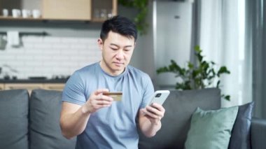 Asyalı adam evdeki modern akıllı telefon ya da cep telefonunu kullanarak online ödeme yapıyor. Kanepede oturuyor ve gülümsüyor. Finans, internetten alışveriş. Mutlu erkek otel, gezi ya da seyahat rezervasyonu 