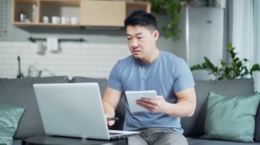 Genç Asyalı erkek çevrimiçi derslerle evde üniversiteye gidiyor. Uzaktan eğitim. E- öğrenim için dizüstü bilgisayar kullanan bir adam danışma videosu için tavsiye ister, bir deftere yazar.
