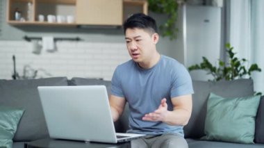 Asyalı özgüvenli bir adam video görüşmesinde uzaktan web kamerası laptopuyla konuşuyor evdeki kanepede oturuyor. Erkek öğretmen ya da öğretim görevlisi Sanal sohbet toplantısı ya da konferansta gelişigüzel iletişim kurar