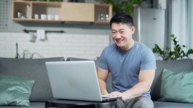 Neşeli, mutlu Asyalı bir adam internetten web kameralı bir bilgisayarla konuşuyor evdeki kanepede oturuyor. Toplantıda gülümseyen erkek el sallaması ya da günlük kıyafetlerle konferans. Sanal sohbet