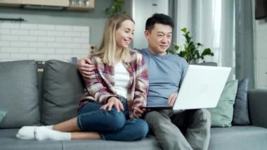 Evli çift uzaktan video görüşmesi yapıyor, evdeki koltukta dizüstü bilgisayar kullanarak oturuyor. Mutlu Asyalı erkek ve kadın el sallıyor ve eğleniyor iletişim kuruyor ve sohbet ediyor.