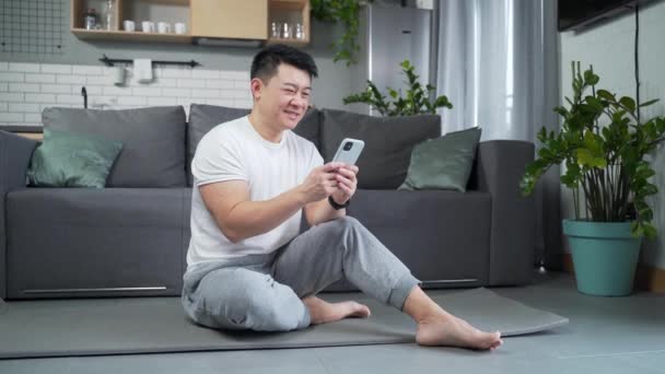 一个亚洲运动员坐在家里的瑜伽垫上 用智能手机 观看视频教程 关于瑜伽的练习 健康男性浏览手机 室内快乐运动员读书 — 图库视频影像