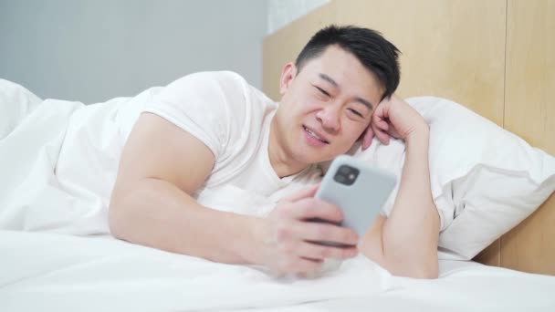 Szczęśliwy Przystojny Azjata Używa Rano Smartfona Leżąc Łóżku Sypialni Środku — Wideo stockowe