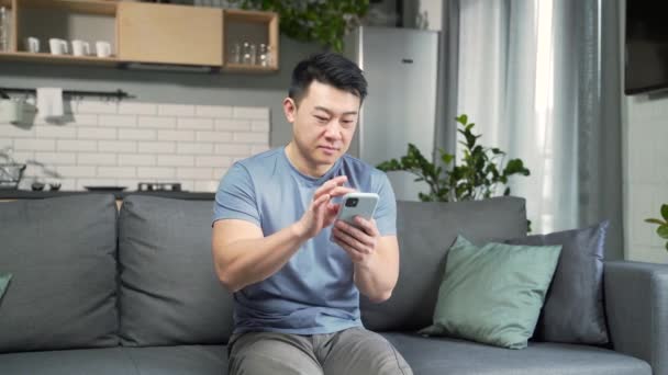 自宅のリビングルームでソファに座っている間にインターネットを閲覧するためにスマートフォンを使用してスクロールするアジアの男 ソファの上でカジュアルな服装で男性をリラックス笑顔幸せな顧客オンライン使用携帯電話 — ストック動画