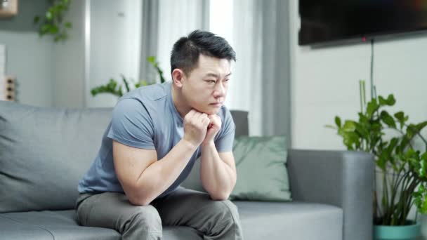 抑郁的亚裔男子想念家乡 可悲的男人坐在沙发上忧心忡忡 他住在客厅里强调家庭或工作中的问题 不治之症 消极思想 — 图库视频影像