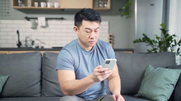 亚洲男人坐在家里的沙发上 用智能手机阅读坏消息 失望地看着智能手机屏幕 对收到的信息感到失望 恼怒的银行贷款通知 — 图库视频影像