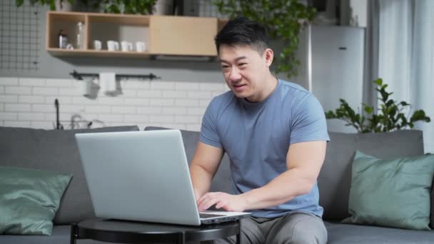 ラップトップの成功を祝うと腕を上げて若いアジアの男 自宅でコンピューターを見ています 興奮した男は ソファの上に座って揺れ リビングルームで良いニュースを読むことに成功し — ストック動画