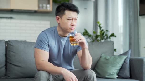 喝多了的亚洲人坐在家里痛痛快快地喝酒 成瘾成性的观念闷闷不乐的男人坐在沙发上 — 图库视频影像