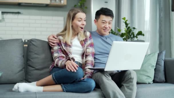 幸せな若い家族の妻と夫は自宅でソファの上に座っている電子メールの使用ノートパソコンは良いニュースを読むオンライン宝くじ受賞者は結果を受け取ります 販売割引 低価格興奮クライアントアジアのカップル喜んで — ストック動画