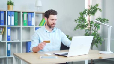 Mutlu, gülümseyen, yetişkin bir adam online alışveriş yapıyor ya da e-alışveriş yapıyor. Çevrimiçi çalışan girişimci, hizmet için ödeme yapıyor. Modern ofisteki dizüstü bilgisayarında kredi kartıyla oturuyor.