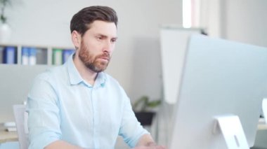 Odaklanmış yakışıklı sakallı iş adamı ofisteki bilgisayar monitörüne ya da laptopa bakıyor. Düşünceli bir girişimci, akıllı tesadüfi bir şekilde kapalı alanda arama yapıyor.