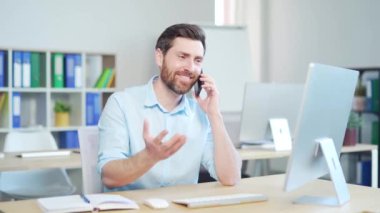 Neşeli ofis çalışanı, çalışan girişimci cep telefonuyla mutlu bir şekilde konuşuyor. İş yerindeki bir bilgisayar masasında oturmak. Kapalı alanda iletişim ve sohbet. İyi habere gül. Gülümse.