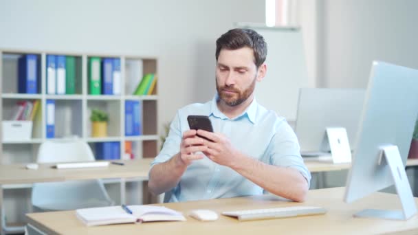 快乐英俊的上班族坐在办公室的电脑桌前 用手机 一个快乐的大胡子商人企业家的雇员 浏览智能手机读消息聊天交流 — 图库视频影像