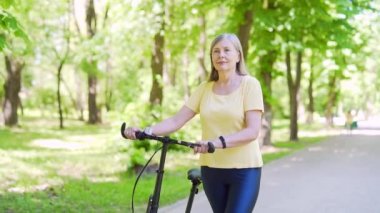 Şehir parkında bisiklet süren olgun, modern, mutlu bir kadın. Yaşlı kadın sağlıklı yaşam tarzı, bisikletle emeklilikte boş zaman. Açık havada, doğada temiz havada yürüyor.
