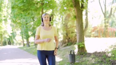 Şehir merkezindeki yaz parkında koşan yaşlı, gri saçlı bir kadın. Olgun, yaşlı bir fitness kadını kulaklıkla koşuyor ve müzik dinliyor. Yaşlı spor koşucusu Kardiyo