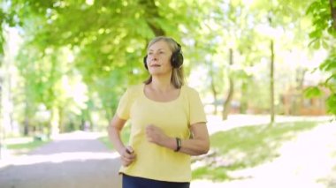 Şehir merkezindeki yaz parkında koşan yaşlı, gri saçlı bir kadın. Olgun, yaşlı bir fitness kadını kulaklıkla koşuyor ve müzik dinliyor. Yaşlı spor koşucusu Kardiyo