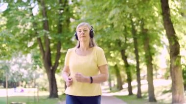 Şehir merkezindeki yaz parkında koşan yaşlı, gri saçlı bir kadın. Olgun, yaşlı bir fitness kadını kulaklıkla koşuyor ve müzik dinliyor. Yaşlı spor koşucusu Kardiyo
