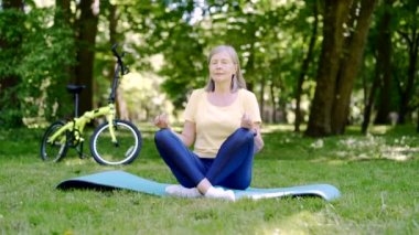 Aktif Gri Saçlı Kıdemli Fitness Kadını yoga pozisyonunda yoga yapıyor. Şehir parkında çimlerin üzerinde yoga minderinin üzerinde oturuyor. Olgun kadın yaşlılar sağlıklı egzersiz yaşam tarzı aktivite sporları