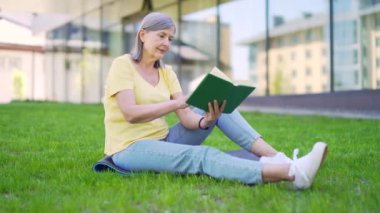 Şehir parkındaki çimlerin üzerinde oturan çayır üzerine kitap okuyan son sınıf öğrencisi bir kadın. Açık havada. Modern mutlu olgun dişi boş zamanlarını doğada geçirir. Dinlen ve temiz hava al. Tasasız yaşam tarzı