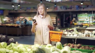 Yaşlı, olgun bir kadın alıcı akıllı telefondan alışveriş listesini kontrol ediyor, süpermarketten alışveriş yapıyor. Modern yaşlı, gri saçlı, emekli kadın alışverişçi cep telefonu kullanıyor.