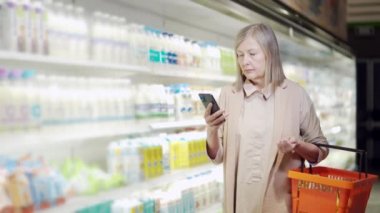 Yaşlı, olgun bir kadın alıcı akıllı telefon kullanarak alışveriş listesini kontrol ediyor, süpermarketten alışveriş yapıyor. Modern yaşlı, gri saçlı, emekli kadın alışverişçi cep telefonu kullanıyor.