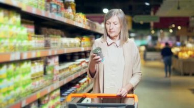 Yetişkin bir kadın markette alışveriş yaparken yiyecek ürünlerini karşılaştırıyor. Emekli yaşlı kadın raflardan mal seçiyor süpermarkette elinde tutuyor, piyasadaki kompozisyonu inceliyor.