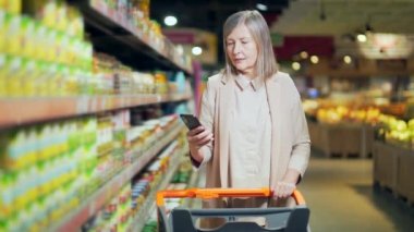 Yaşlı, olgun bir kadın alıcı akıllı telefon kullanarak alışveriş listesini kontrol ediyor, süpermarketten alışveriş yapıyor. Modern yaşlı, gri saçlı, emekli kadın alışverişçi cep telefonu kullanıyor.