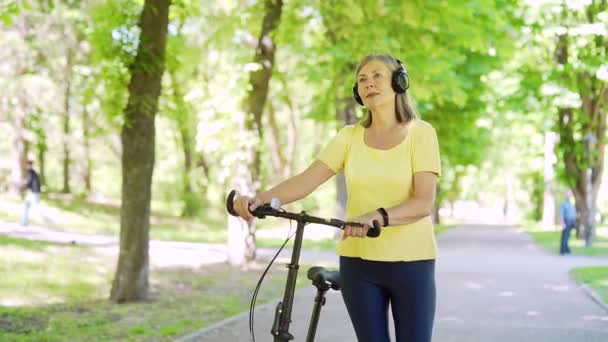 快乐的年长的 成熟的 活跃的现代女性 带着耳机骑自行车在城市公园里散步 欣赏和听音乐 老年女性健康的生活方式和骑自行车退休后的闲暇时间 — 图库视频影像