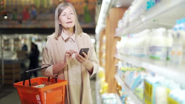 お年寄りのおばあさんスマホで買い物リストを確認したり スーパーの食料品店で買い物をしたり 現代の白髪年金受給者女性の買い物客は 携帯電話を使用して選択します — ストック動画