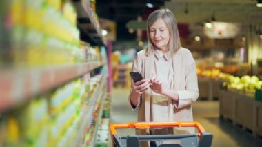 Yaşlı, olgun bir kadın alıcı akıllı telefon kullanarak alışveriş listesini kontrol ediyor, süpermarketten alışveriş yapıyor. Modern yaşlı, gri saçlı, emekli kadın alışverişçi cep telefonu kullanıyor.