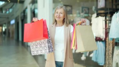 Alışveriş merkezinde kameraya bakıp alışveriş torbalarını gösteren son sınıf bir bayan müşterinin portresi. Mutlu olgun yaşlı kadın satın aldıktan sonra hediyelerle hava atar. Gülümse. Mağazanın memnun, yaşlı müşterisi.