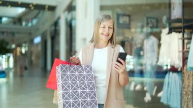 Alışveriş merkezindeki son sınıf bayan müşterinin portresi akıllı telefon kullanıyor. Alışveriş çantalı akıllı cep telefonu uygulaması kullanarak taramalar. Satın aldıktan sonra mutlu olgun bir kadın. Gülümse. Mağazanın memnun yaşlı müşterisi