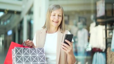 Alışveriş merkezindeki son sınıf bayan müşterinin portresi akıllı telefon kullanıyor. Alışveriş çantalı akıllı cep telefonu uygulaması kullanarak taramalar. Satın aldıktan sonra mutlu olgun bir kadın. Gülümse. Mağazanın memnun yaşlı müşterisi