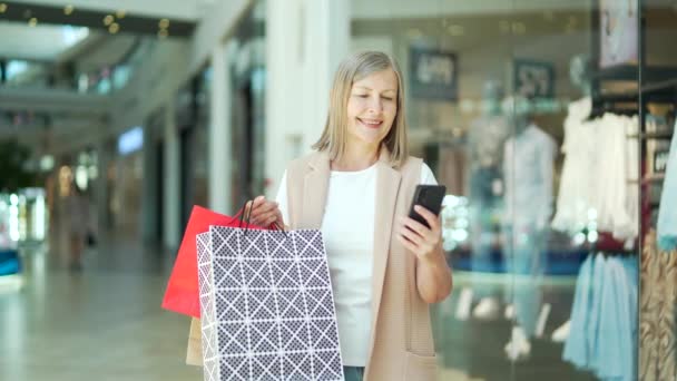 モール内のポートレートシニア女性買い物客はスマートフォンを使用しています 買い物袋付きのアプリスマートフォンで閲覧できます 購入後の幸せな成熟した女性 笑顔だ 店舗の満足長老顧客 — ストック動画
