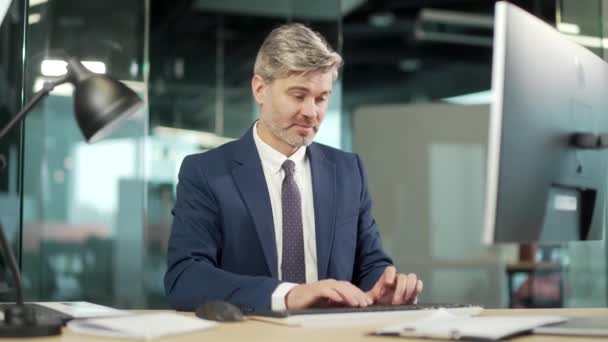 成功的成熟商人 穿着合身的工作服 坐在办公桌前 笔记本电脑在办公室 在工作场所电脑上工作的一个自信的白发男性的画像 笑着看着相机的大拇指向上看 — 图库视频影像