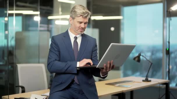 ポートレート 成熟したひげカメラを見てスーツの成功したビジネスマン 現代のオフィスに立っている間 手にノートパソコンのコンピュータで作業 灰色の髪の自信のある起業家男性が職場で — ストック動画
