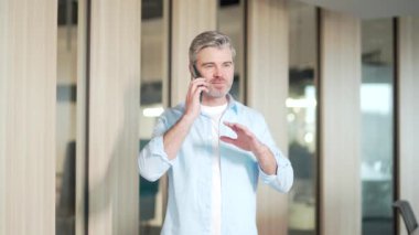 Gri sakallı mutlu iş adamı gündelik giysiler içinde konuşuyor, cep telefonuyla iletişim kuruyor modern ofiste ayakta duruyor. Üst düzey çalışan, akıllı telefonlardan konuşur. gülümsüyor 