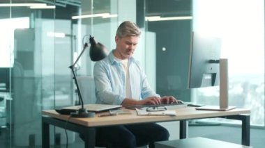 Masadaki modern cam ofiste bilgisayar işyerinde çalışan gri saçlı, kendine güvenen, sıradan giyinmiş bir iş adamı. Yetişkin, yaşlı, sakallı, iş yerinde çalışan iş adamı.