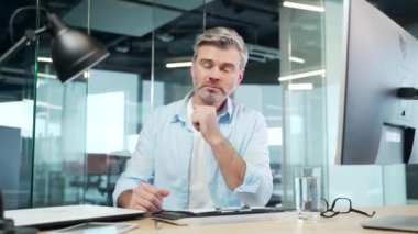 Olgun sakallı adam internetteki seminer toplantılarını, konferansları ya da konferansları kameraya uzaktan bakan eğitimleri dinliyor. Yaşlı gri saçlı, video başında çalışan bir erkek ofis masasında oturuyor.