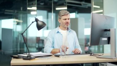 Olgun bir erkek ofis çalışanı çalışma yerinde masa başında bilgisayar başında çalışıyor. İş adamı bardaktan temiz su içer. Gri saçlı, sakallı, yakışıklı iş adamı susuzluğunu gideriyor.