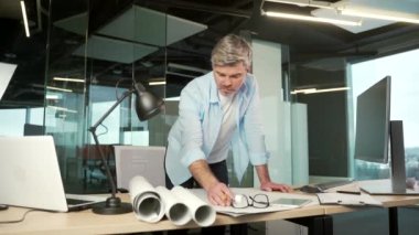 Olgun yaratıcı tasarımcı, ofiste çalışan bir mimar çizimlerle dolu bir bilgisayar masası. Yakışıklı son sınıf öğrencisi iş yerinde dikilip ev planı çiziyor.